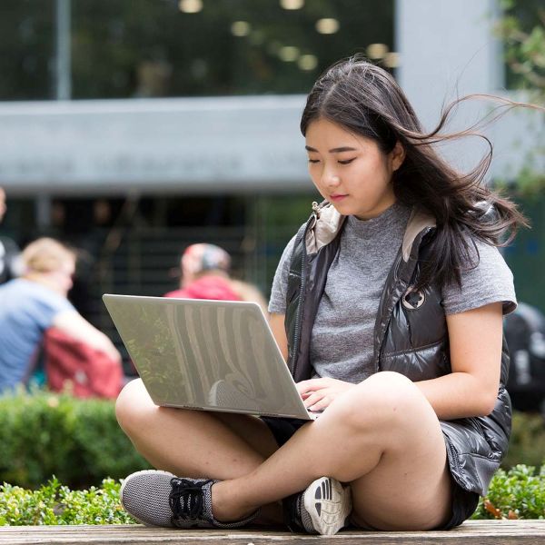 Student sitting crosslegged outside working on their open laptop.