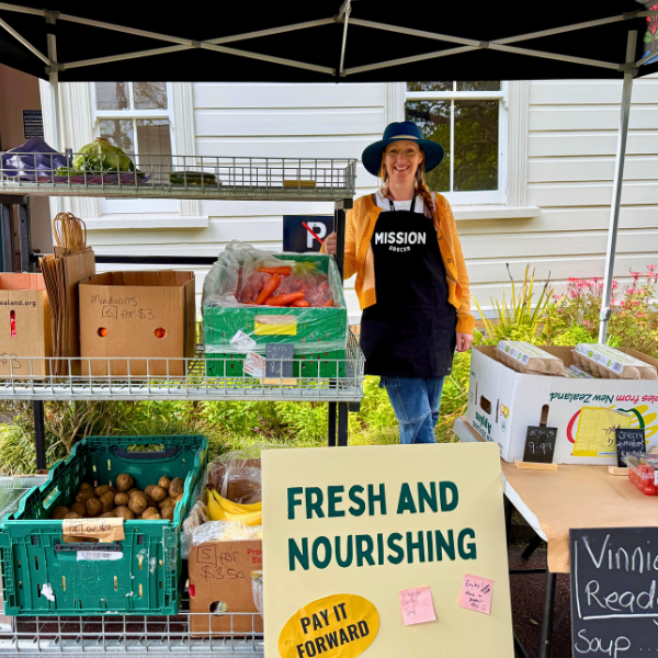 Fresh produce stall with boxes of vegetables and fruit, a volunteer in a black apron and hat.